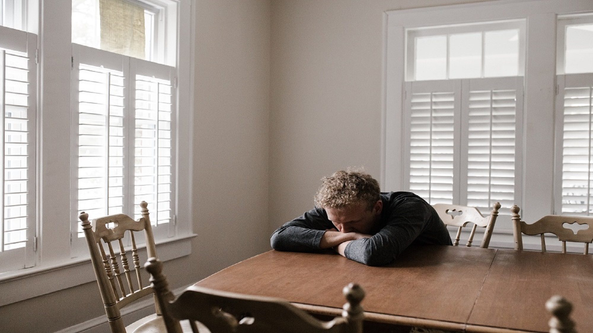 Man alone in dining room with his head against the table and arms crossed to his head