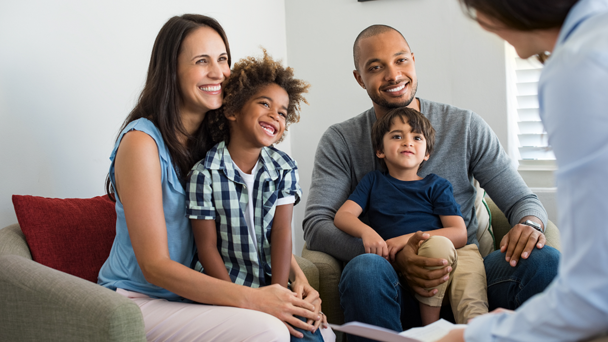 Smiling family sitting on chairs, with young boy sitting on father's lap and young girl sitting on mother's lap, all facing therapist who is holding piece of paper