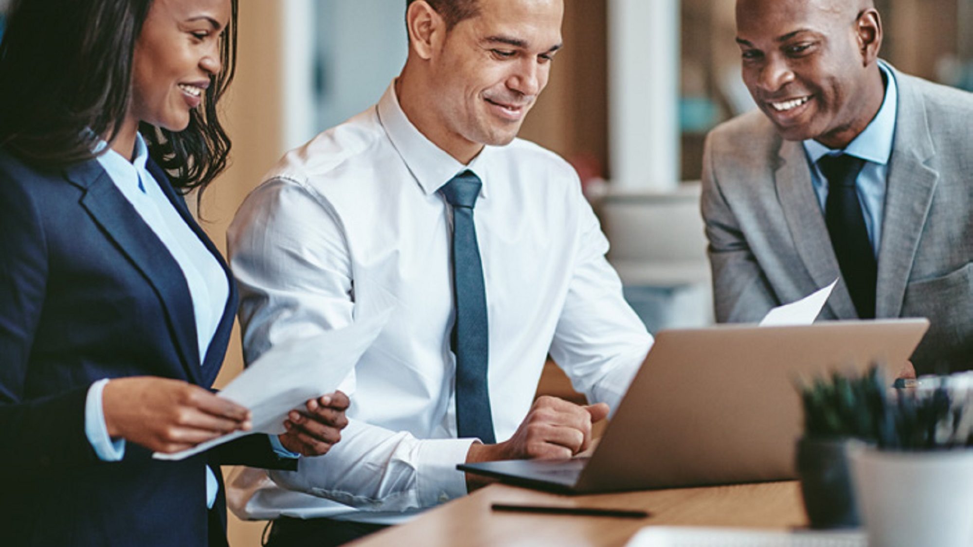 two businessmen and woman in an office looking at a laptop on a desk with a plant